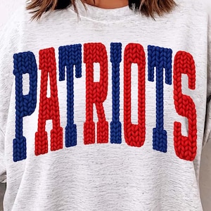 May include: A light gray sweatshirt with the word "PATRIOTS" in large, textured, red and blue letters across the front. The sweatshirt has long sleeves and a crew neck.