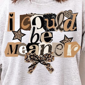 May include: A light gray sweatshirt with the text "I could be meaner" in a collage of fonts and patterns. The design includes a bow, stars, and a heart. The sweatshirt is a casual, comfortable style.