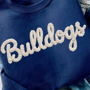 May include: Navy blue sweatshirt with the word "Bulldogs" in a cream-colored, rope-like font. The sweatshirt is folded on top of a pair of blue jeans. The image is well-lit and shows the details of the fabric.
