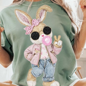 Può includere: T-shirt verde salvia con un design a forma di coniglio dei cartoni animati. Il coniglio indossa occhiali da sole, una giacca rosa e jeans blu, soffia una bolla di gomma da masticare e fa il segno della pace. Il coniglio ha un fiocco rosa e indossa occhiali da sole.
