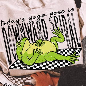 Może przedstawiać: Kremowa bluza z napisem "today's yoga pose is DOWNWARD SPIRAL" i kreskówkową żabą na czarno-białej szachownicy. Widoczna jest również kurtka dżinsowa i para szachownicowych trampek.