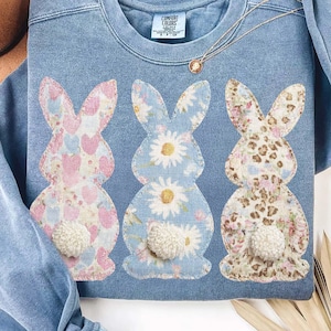 Può includere: Una felpa blu girocollo con tre coniglietti applicati con disegni floreali e fantasia. Ogni coniglietto ha una soffice coda a pompon bianca. Una collana d'oro con un ciondolo è drappeggiata sulla felpa.