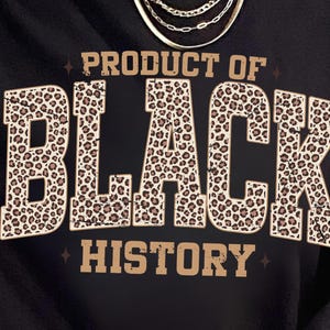 May include: Black sweatshirt with the text "PRODUCT OF BLACK HISTORY" in a bold, leopard print font. The sweatshirt has a crew neck and is worn with a gold necklace. The design is centered on the front of the shirt.