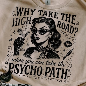 May include: A beige t-shirt featuring a graphic of a woman in sunglasses holding a cocktail, with the text "WHY TAKE THE HIGH ROAD? when you can take the PSYCHO PATH." A black and white checkered fanny pack and sunglasses are also visible.