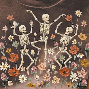 May include: A dark brown crewneck sweatshirt featuring a graphic print of three dancing skeletons surrounded by various colourful wildflowers in shades of orange, pink, white, and green. The design is centred on the front of the garment.