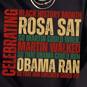 May include: Black sweatshirt with text celebrating Black History Month. The text includes "ROSA SAT, SO MARTIN COULD WALK, MARTIN WALKED, SO THAT OBAMA COULD RUN, OBAMA RAN, SO THAT OUR CHILDREN COULD FLY."