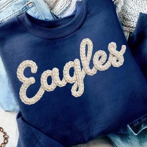 Peut inclure: Un sweat-shirt bleu marine avec le mot "Eagles" brodé dans une police texturée de couleur crème. Le sweat-shirt est posé sur un jean bleu clair. Un bracelet chaîne de couleur or est visible.
