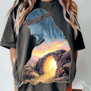 Könnte beinhalten: Dunkelgraues T-Shirt mit einer Silhouette von Jesus mit einer Dornenkrone, gefüllt mit einer Sonnenuntergangs- und Höhlenszene. Der Text "digital download" ist sichtbar. Teil der Infinite Pixel Studio Kollektion.