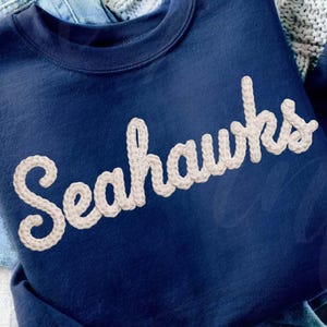 Puede incluir: Una sudadera azul marino con la palabra "Seahawks" bordada en escritura color crema en el pecho. La sudadera está doblada sobre un par de vaqueros azules. La imagen está bien iluminada y muestra los detalles de la tela.