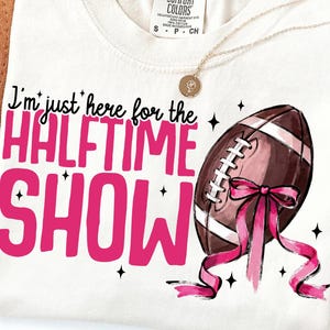 May include: Off-white t-shirt featuring the text "I'm just here for the Halftime Show" in pink. A brown American football with pink ribbon is on the right. A gold necklace is draped across the shirt.