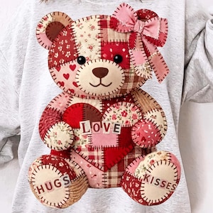 Può includere: Una felpa grigio chiaro con un design a orsetto patchwork. L'orso ha un cuore con la parola "LOVE" e le parole "HUGS" e "KISSES". L'orso è fatto di tessuto rosso, rosa e beige con un fiocco rosa.