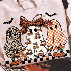 May include: A cream-colored sweatshirt featuring three ghost characters with Halloween-themed designs. One ghost has a leopard print, another has a bow, and the third has a checkered pattern. The design includes pumpkins, bats, and stars. The words "digital download" are visible.