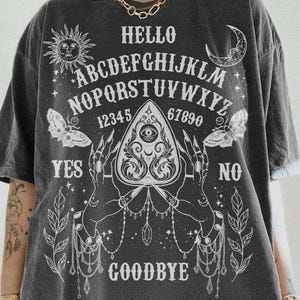 May include: Charcoal grey t-shirt featuring a white Ouija board graphic. The design includes the alphabet, numbers, sun, moon, butterflies, and the words "Hello", "Yes", "No", and "Goodbye".