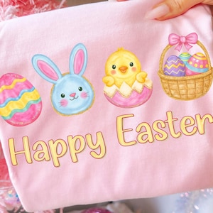 May include: A light pink t-shirt featuring Easter-themed illustrations. The design includes a decorated egg, a blue bunny, a yellow chick hatching from an egg, and a basket of decorated eggs. The words "Happy Easter" are printed in yellow.