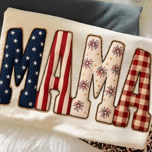 Puede incluir: Sudadera color crema con la palabra "MAMA" en patrones patrióticos. Las letras están delineadas con un borde marrón en forma de cuerda. La "M" es azul con estrellas blancas, la "A" tiene rayas rojas y blancas, y las otras letras tienen patrones de fuegos artificiales y cuadros.