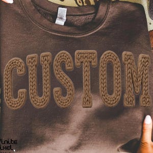 May include: A brown sweatshirt with the word "CUSTOM" in a textured, knitted style across the front. The sweatshirt is held up by two hands. The text is in a lighter shade of brown than the sweatshirt.