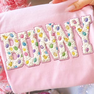 May include: A pale pink t-shirt featuring the word "BUNNY" embroidered in white, adorned with colourful Easter eggs and yellow chicks. The shirt is held up by a hand, with a pink Christmas tree in the background.