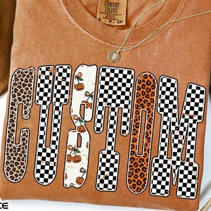 Peut inclure: Un t-shirt Comfort Colors orange brûlé avec le mot "CUSTOM" en police dégoulinante. Les lettres présentent des motifs léopard, damier et citrouille. Un collier en or et un chapeau marron sont également visibles.