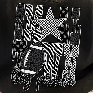May include: Black sweatshirt with a graphic design featuring a football, star, and the words "Small Town Big Pride." The design incorporates zebra stripes, leopard print, and a checkered pattern. The text is in a handwritten style.