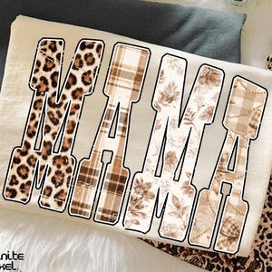 Può includere: T-shirt color crema con la scritta "MAMA" a caratteri grandi e in grassetto. Ogni lettera presenta un motivo diverso: stampa leopardata, scozzese, floreale e un design a fantasia. La maglietta è piegata su una superficie grigia.