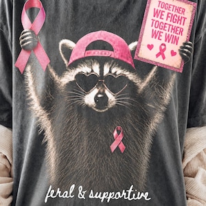Puede incluir: Una camiseta gris carbón con el gráfico de un mapache que lleva una gorra rosa y gafas de sol en forma de corazón. El mapache sostiene un lazo rosa y un cartel que dice TOGETHER WE FIGHT TOGETHER WE WIN. El texto inferior dice feral & supportive.