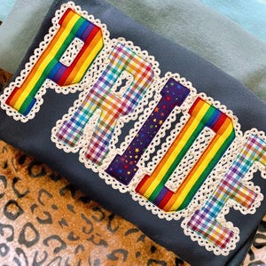 May include: A dark gray t-shirt with the word "PRIDE" appliqued in rainbow colors, plaid, and star patterns, outlined with white lace trim. The shirt is folded, with other folded shirts in the background.
