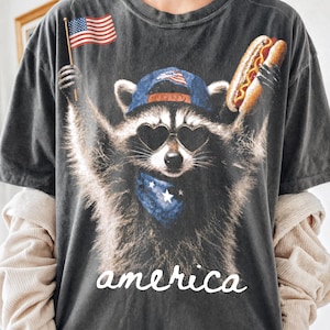 Op de afbeelding: Donkergrijs t-shirt met een wasbeer met een hartvormige zonnebril, een blauwe bandana en een baseballpet met een Amerikaans vlagontwerp. De wasbeer houdt een hotdog en een Amerikaanse vlag vast. Het woord "america" is in witte letters geschreven.