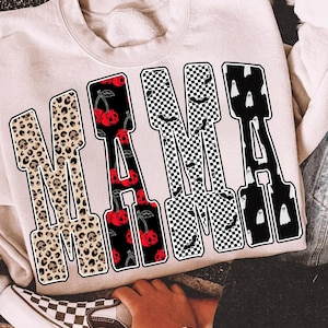 May include: A cream-coloured sweatshirt displaying the word "MAMA" in a bold, patterned design. The letters feature leopard print, cherries, checkered patterns, and ghosts. A denim jacket and a checkered slip-on shoe are also visible.
