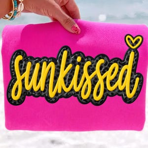 Puede incluir: Una toalla de playa rosa brillante con la palabra "Sunkissed" en escritura amarilla con un fondo de estampado de leopardo negro y gris. Un corazón amarillo está encima de la palabra. La toalla está sostenida por una mano con una pulsera.