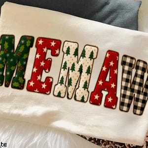 May include: A cream-colored sweatshirt with the word "MEMAW" in large, festive letters. The letters are decorated with Christmas-themed patterns: holly, red stars, Christmas trees, and black and white buffalo check. The sweatshirt is folded on a white surface.
