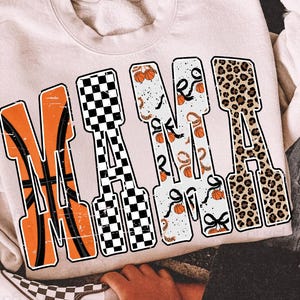 Puede incluir: Sudadera color crema con la palabra "MAMA" en letras grandes y audaces. Las letras est&aacute;n dise&ntilde;adas con varios patrones, incluyendo baloncesto, cuadros y estampado de leopardo. Se ve una chaqueta vaquera con dise&ntilde;os de llamas.