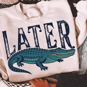 Pode incluir: Uma camisa de moletom bege com a palavra "LATER" em uma fonte desgastada e um gr&aacute;fico de um jacar&eacute; verde.
