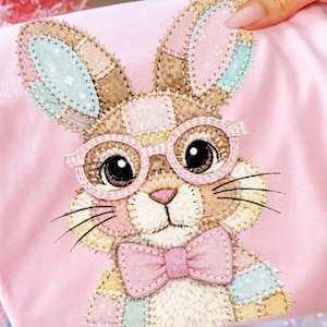 May include: A pink t-shirt featuring a patchwork bunny wearing glasses and a bow tie. The bunny has brown, pink, and blue accents. The t-shirt is held up by a hand, with a pink Christmas tree and ornaments in the background.
