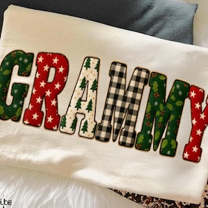 May include: Cream-colored sweatshirt with the word "GRAMMY" in a festive, patchwork design. The letters feature patterns like red stars, green holly, Christmas trees, and black and white buffalo check. The sweatshirt is folded on a white, fluffy surface.