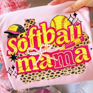 Può includere: Maglietta rosa chiaro con le parole "Softball Mama" in giallo e rosso, insieme a una grafica di baseball, casco e mazza. Il design include uno sfondo con stampa leopardata e un fiocco di perle.