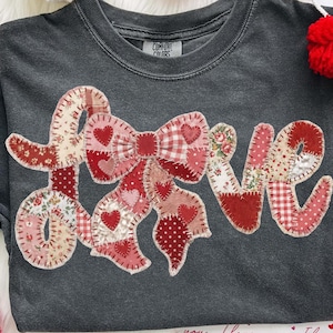 Könnte beinhalten: Ein dunkelgraues T-Shirt mit dem Wort "LOVE", das in einem Patchwork-Design aus roten und rosa Stoffen appliziert ist. Die Schleife und die Buchstaben weisen Herzen und Blumenmuster auf. Das Shirt ist von roten Herzdekorationen und Pompons umgeben.