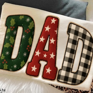 May include: Cream-coloured sweatshirt featuring the word "DAD" in festive lettering. The "D" is green with holly and berries, the "A" is red with white stars, and the final "D" is black and white buffalo plaid.