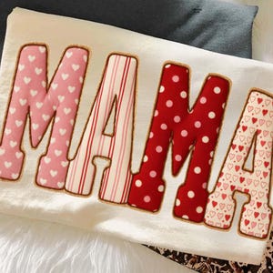May include: Cream-colored t-shirt with the word "MAMA" in large, colorful letters. The letters are outlined in a brown color and filled with patterns like hearts, stripes, and polka dots. The shirt is laying on a white, fluffy surface.