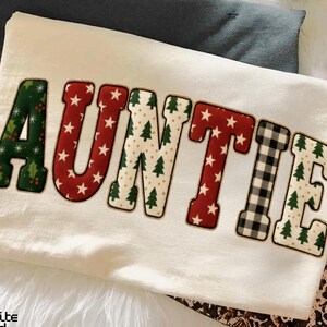 Könnte beinhalten: Cremefarbenes Sweatshirt mit dem Wort "AUNTIE" in einem festlichen Patchwork-Design. Die Buchstaben zeigen Muster aus roten Sternen, grünem Stechpalmen, Weihnachtsbäumen und schwarz-weißem Büffelkaromuster, ideal für die Weihnachtszeit.