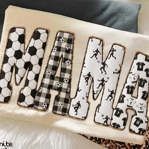 May include: A cream-coloured decorative pillow with the word "MAMA" in large, bold letters. The letters are decorated with football patterns, football player silhouettes, and football jerseys. The pillow is on a white surface.