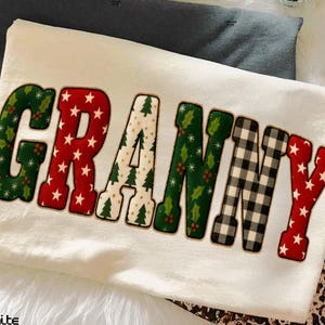 May include: A cream-colored t-shirt with the word "GRANNY" in a festive, decorative font. The letters feature holiday patterns: red stars, green holly, white Christmas trees, and black and white plaid. The shirt is folded on a white surface.