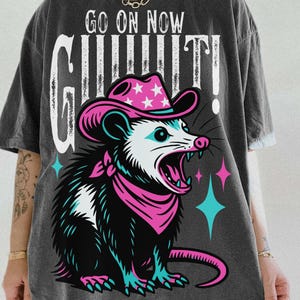 May include: Dark gray t-shirt featuring a cartoon opossum wearing a pink cowboy hat and bandana, with the text "GO ON NOW GUILTY!". The opossum has an open mouth and is surrounded by pink and teal stars.