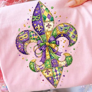 Peut inclure: Un t-shirt rose clair orné d'un motif de fleur de lys coloré. La fleur de lys est décorée de motifs arlequin violets, verts et dorés, de masques et d'un nœud. Le design est agrémenté de petits détails en forme de confettis.