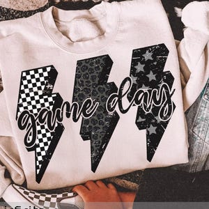 Peut inclure: Sweat-shirt crème avec l'inscription "Game Day" en écriture cursive. Le motif est composé de trois éclairs avec différents motifs : damier, imprimé léopard et étoiles. Une veste en jean avec des détails de flammes est visible.
