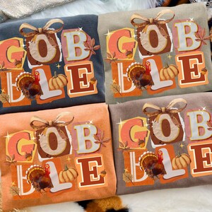 May include: Four crew neck t-shirts in various colors, each featuring the word "GOBBLE" in a fall-themed design. The design includes a turkey, pumpkins, and autumn leaves. The shirts are in shades of gray, tan, orange, and brown.