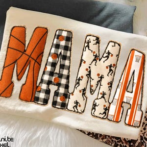 May include: Cream-colored sweatshirt with the word "MAMA" in large, decorative letters. The "M" is orange with basketball lines, the first "A" is black and white checkered, the second "M" has basketball player silhouettes, and the last "A" is orange with basketball net stripes.