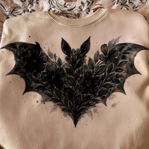 May include: A cream-coloured sweatshirt with a black bat and floral design. The bat's wings are outstretched, and the design includes flowers and leaves. The words "infinite pixel" and "digital download" are visible.