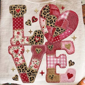 May include: A cream-colored sweatshirt with the word "LOVE" appliqued on the front. The letters are made of various fabrics, including leopard print, floral, and polka dots, with heart and star accents. The sweatshirt is folded.