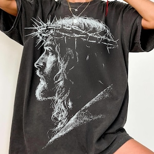 Puede incluir: Camiseta gris oscuro con un gráfico en blanco y negro de un hombre con una corona de espinas. La imagen está centrada en la parte delantera. La camiseta tiene mangas cortas y un corte holgado.