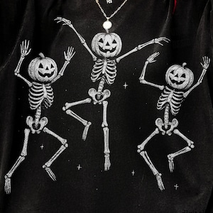 May include: Black t-shirt featuring a Halloween design with three dancing skeletons. Each skeleton has a carved pumpkin for a head and is in a playful pose. The design is in white, creating a contrast against the dark fabric.